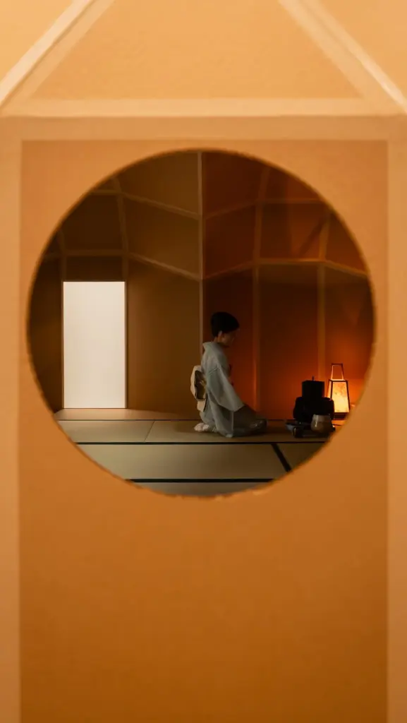 Person in a pale kimono kneeling beside tea utensils in a warmly lit room