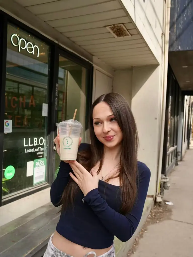 Woman holding a clear ICHA TEA cup with a pale iced drink outside a storefront