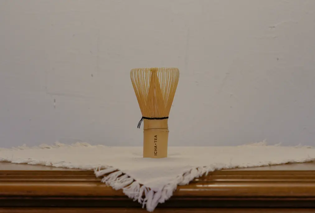 Pale bamboo matcha whisk with fine curved tines and dark thread binding, standing upright on a white fringed cloth