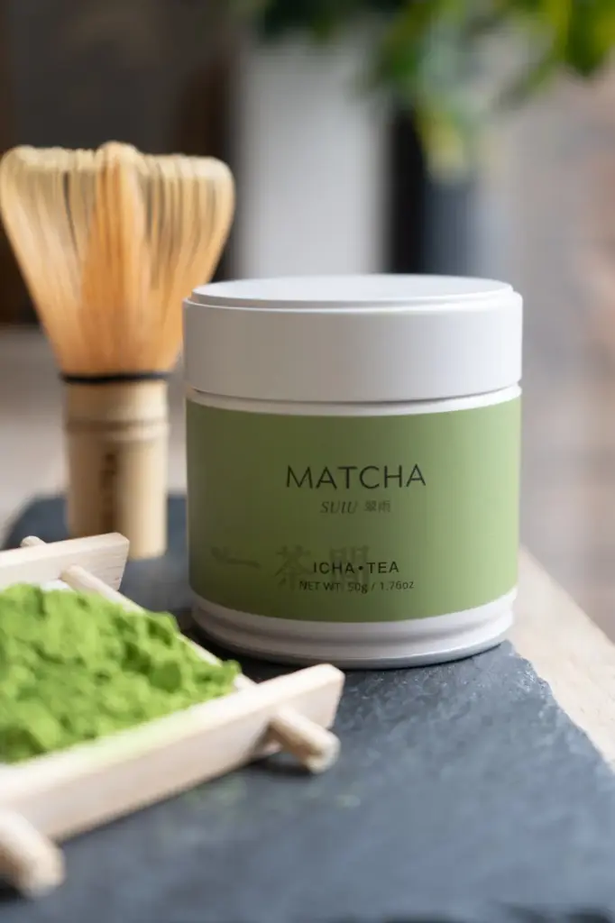 White ICHA TEA Matcha Suiu 50g tin with green label on slate, beside a bamboo whisk and a wooden tray of bright green matcha powder