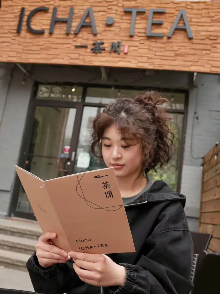 Woman reading an ICHA TEA menu beneath the storefront sign