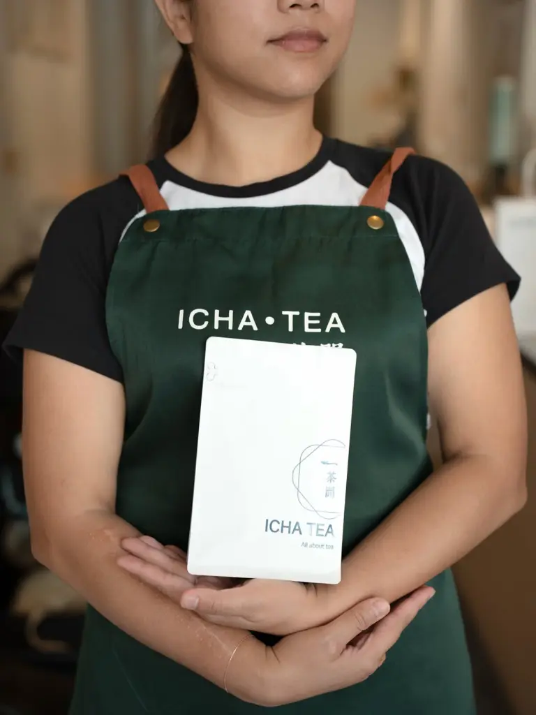 Staff member in a green ICHA TEA apron holding a white branded pouch