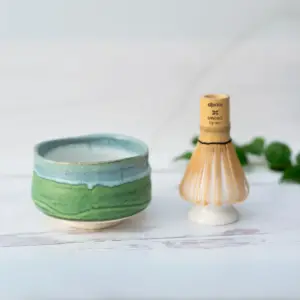 Green and blue glazed matcha bowl beside a bamboo whisk resting on a white ceramic whisk holder, on white wood with ivy leaves behind