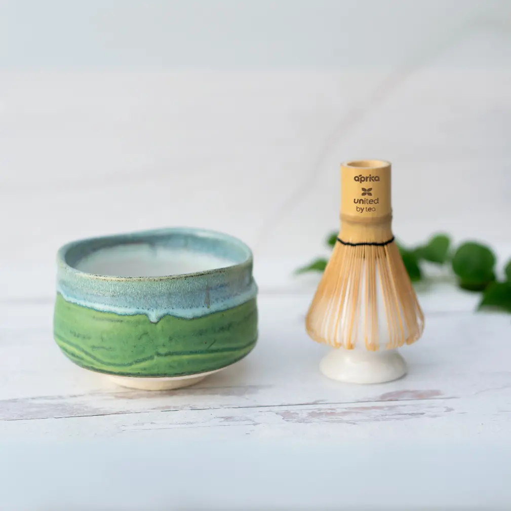 Green and blue glazed matcha bowl beside a bamboo whisk resting on a white ceramic whisk holder, on white wood with ivy leaves behind