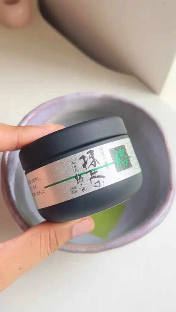 Hand holding a black tea container above a ceramic bowl of green tea