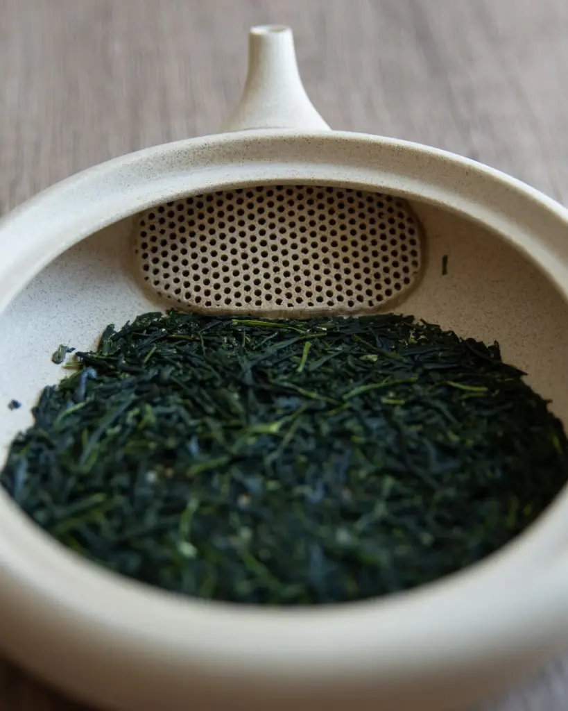 Dark green dry tea leaves inside a beige ceramic teapot with a built-in strainer