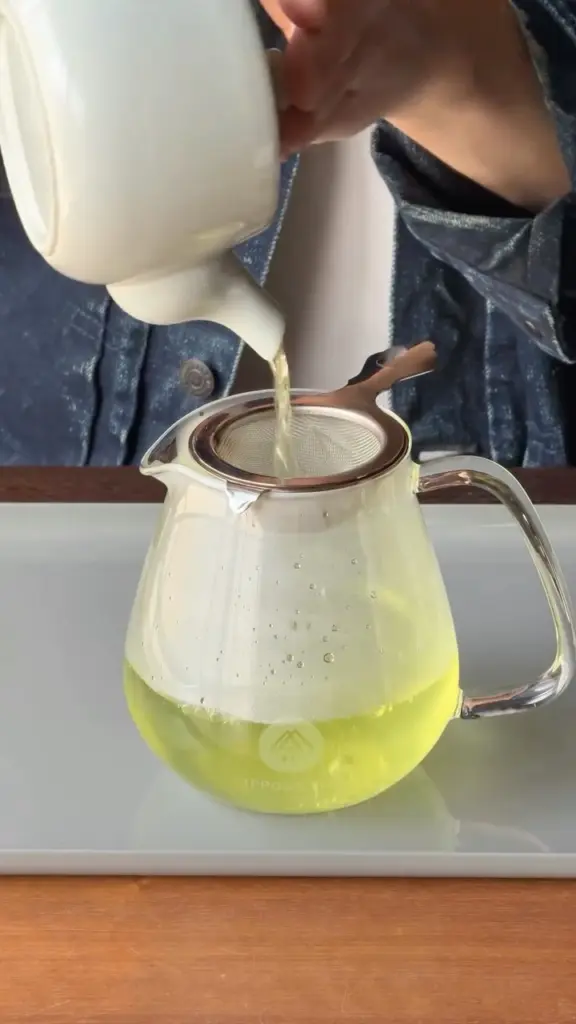 Person pouring pale green tea through a strainer into a glass pitcher