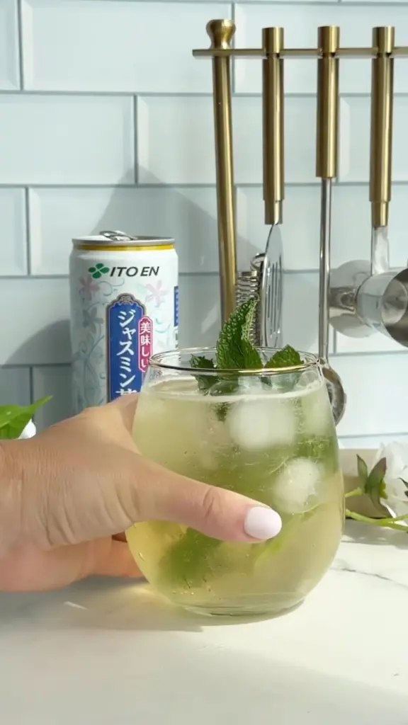 Hand holding a glass of iced green tea garnished with mint beside an Ito En can