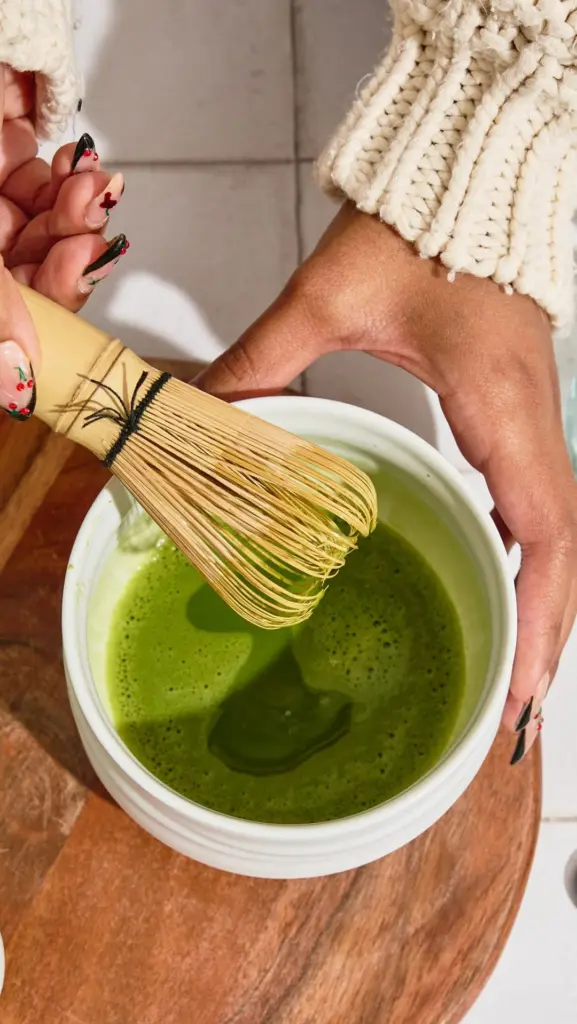 Hands whisking bright green matcha in a white bowl with a bamboo whisk