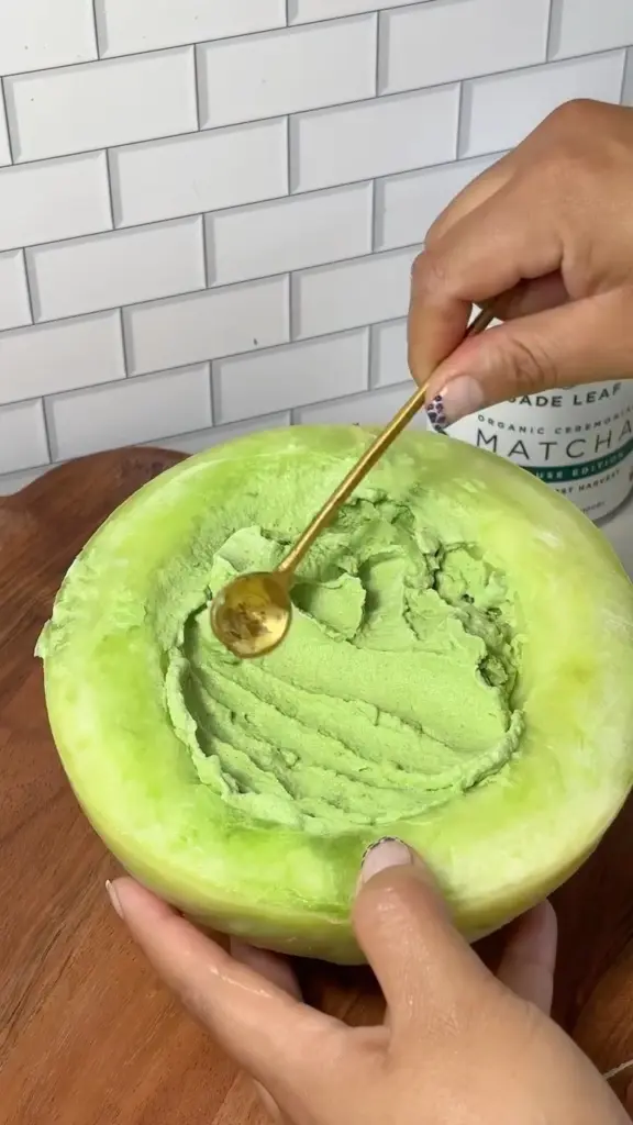 Hands scooping a pale green frozen dessert from a halved melon