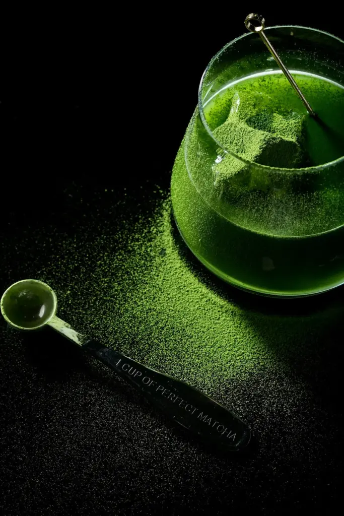 Glass bowl of green matcha powder with two metal measuring spoons on a black surface