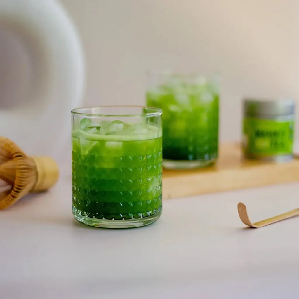 Glass of bright green iced matcha with a bamboo whisk and scoop nearby