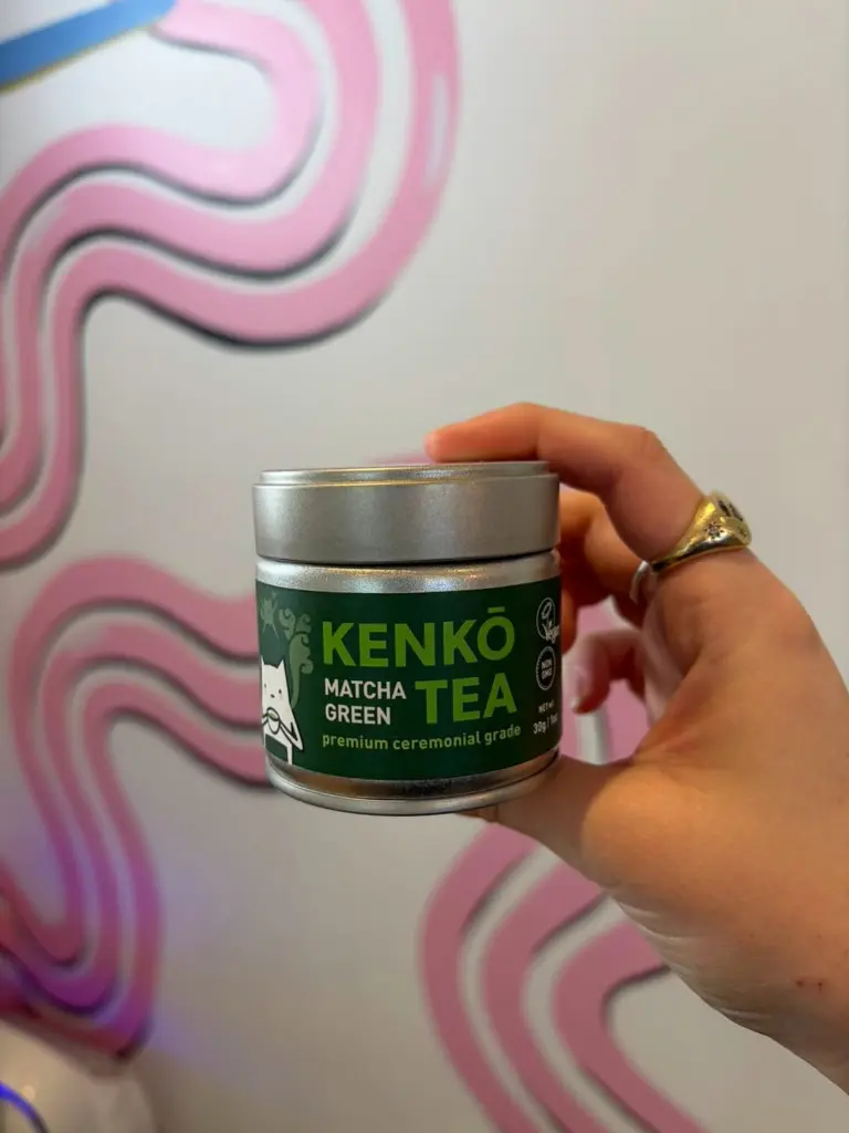 Hand holding a Kenko Tea matcha tin against a pink wavy mural