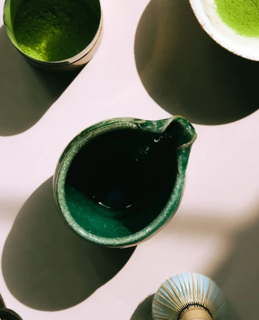 Green ceramic pitcher, matcha containers, and bamboo whisk in sunlight