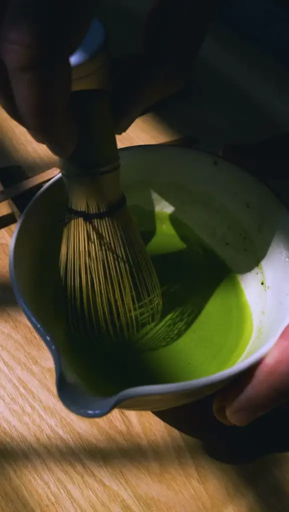 Bamboo whisk stirring bright green matcha in a ceramic bowl