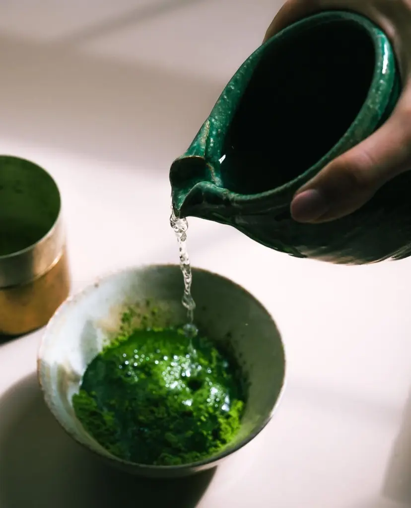 Water pouring from a green ceramic pitcher into a bowl of matcha
