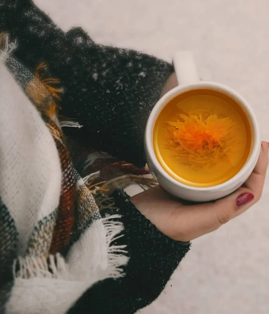 Hand holding a white cup of amber tea with a blooming flower
