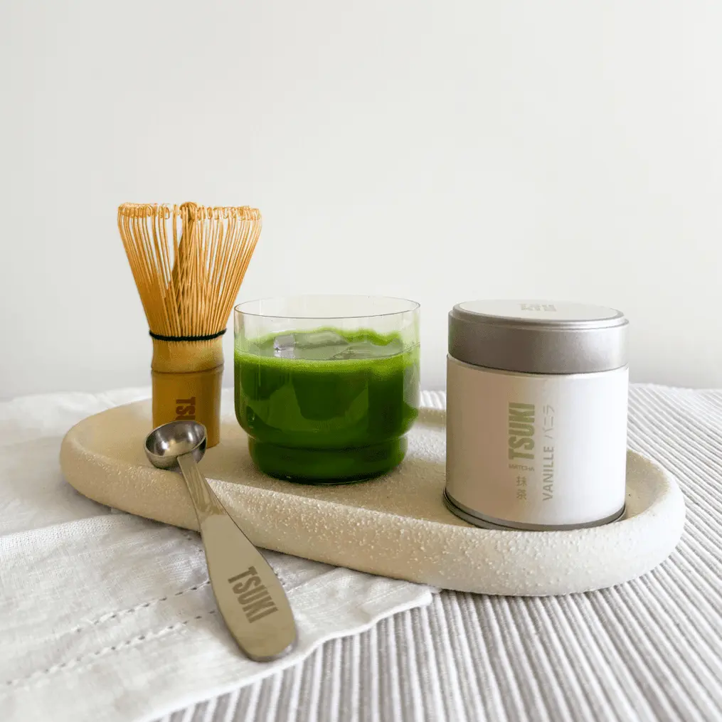 Bamboo whisk, metal scoop, green matcha drink, and Tsuki Vanille tin arranged on a tray