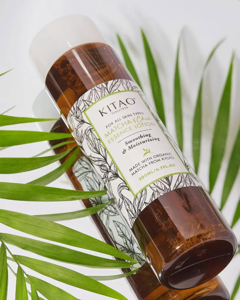 Amber bottle of Kitao Matcha & Chia Essence Lotion among green leaves on a reflective surface