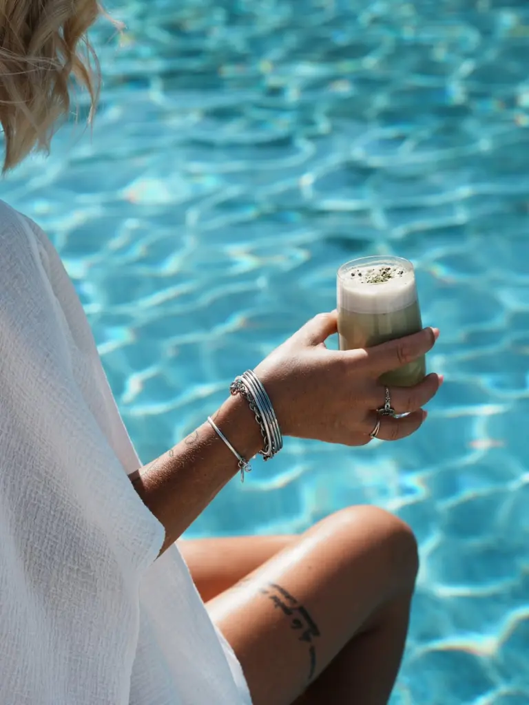 Person holding a foamy green drink beside blue pool water