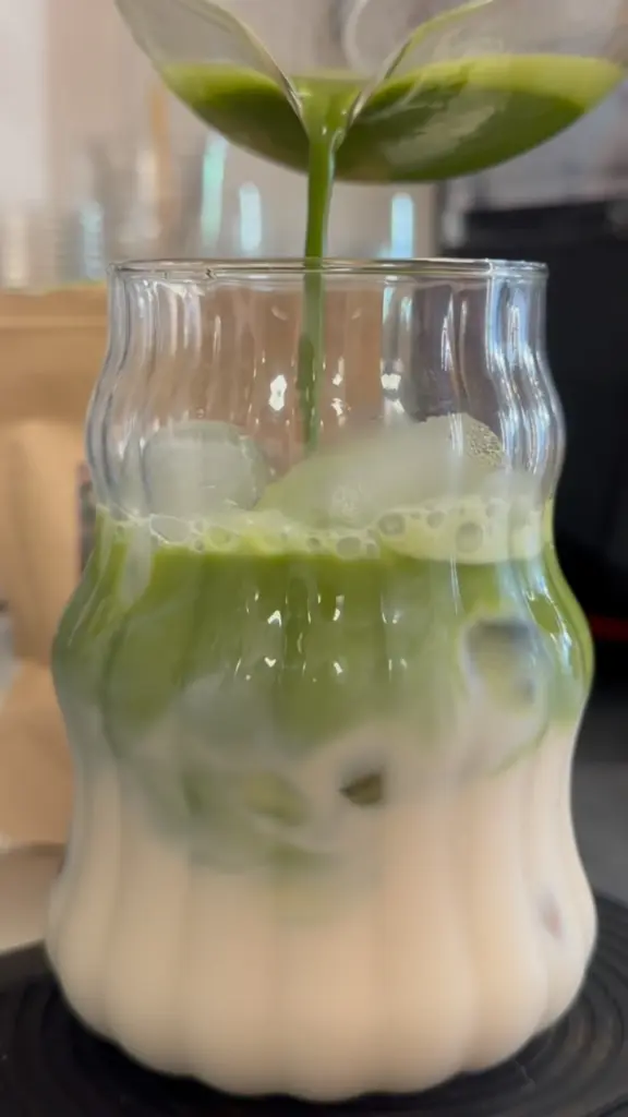Green matcha pouring into a curved glass of iced milk