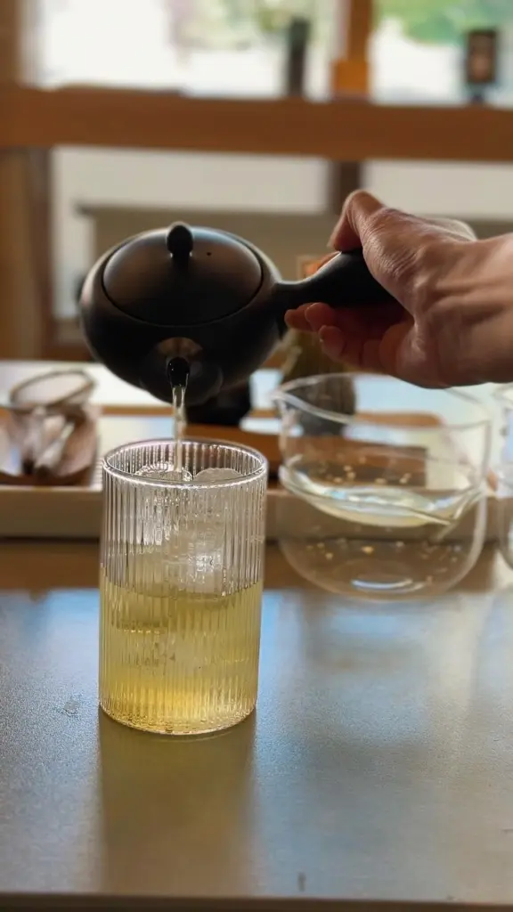 Hand pouring pale green tea from a black teapot into a ribbed glass
