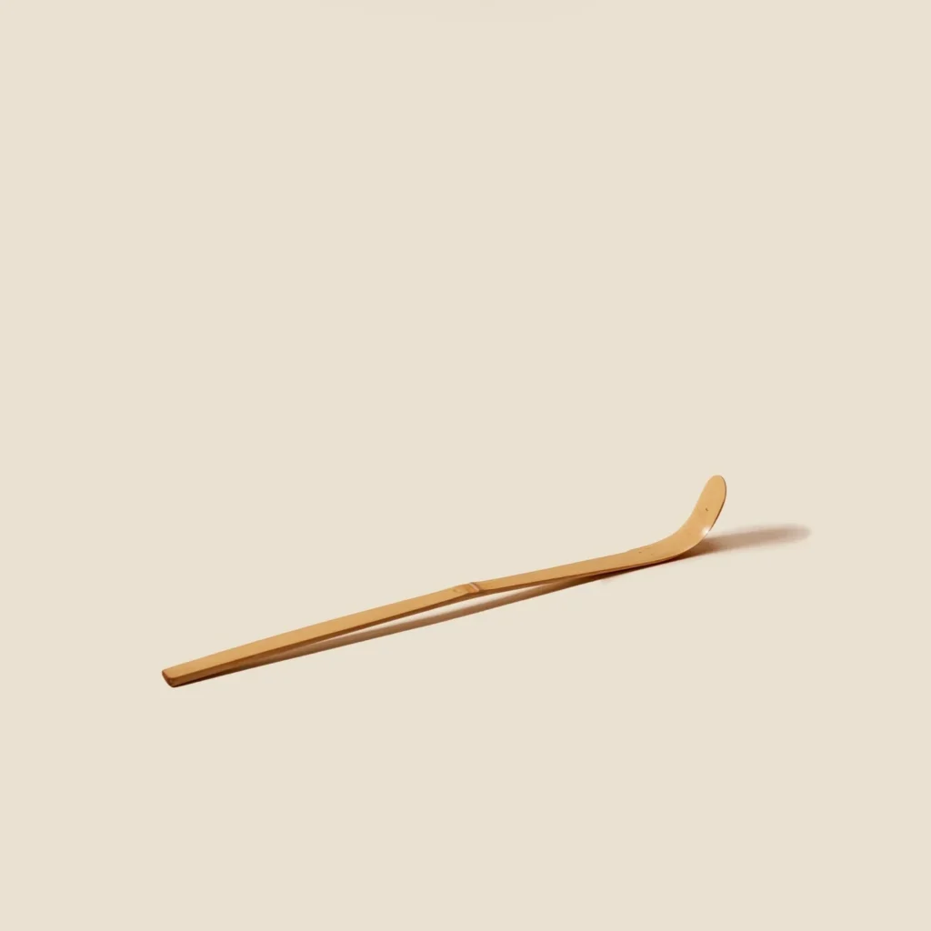 Pale bamboo chashaku tea scoop lying diagonally, curved bowl end raised, on a cream background