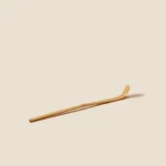 Pale bamboo chashaku tea scoop lying diagonally, curved bowl end raised, on a cream background