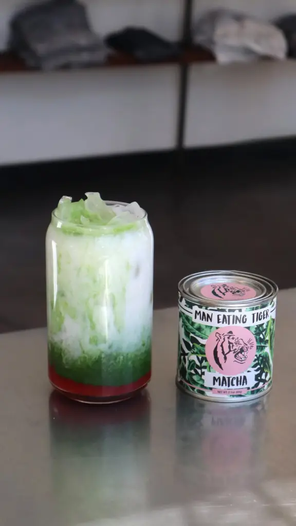 Layered green and white iced drink beside a Man Eating Tiger matcha tin