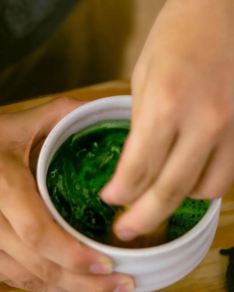 Hands stirring dark green matcha in a white ceramic bowl