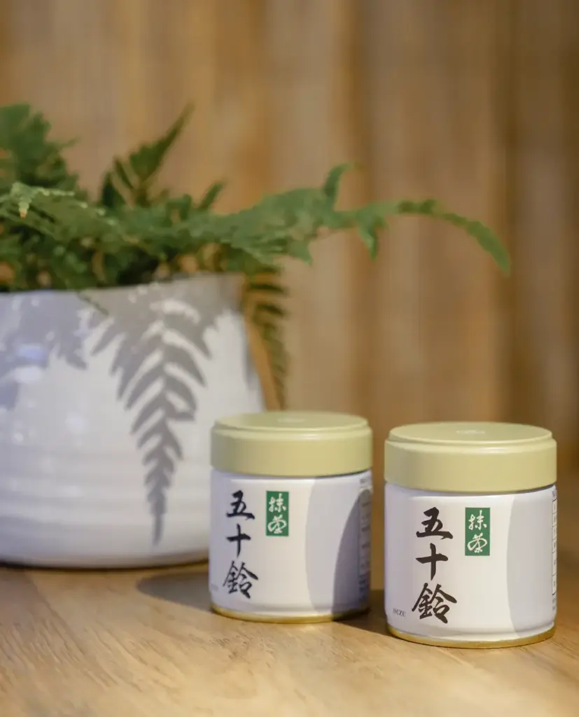 Two white matcha tins labeled 五十鈴 (Isuzu) with pale green lids standing on a wooden table beside a potted fern