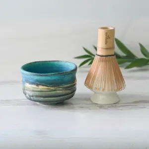 Blue and cream glazed matcha bowl beside a bamboo whisk resting upside down on a white ceramic whisk holder
