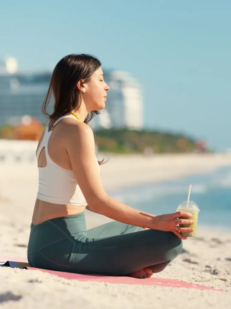 Woman seated cross-legged on a beach mat holding a green drink
