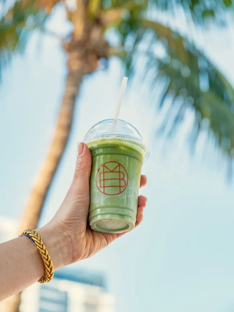 Hand holding a clear cup of green drink beneath a palm tree