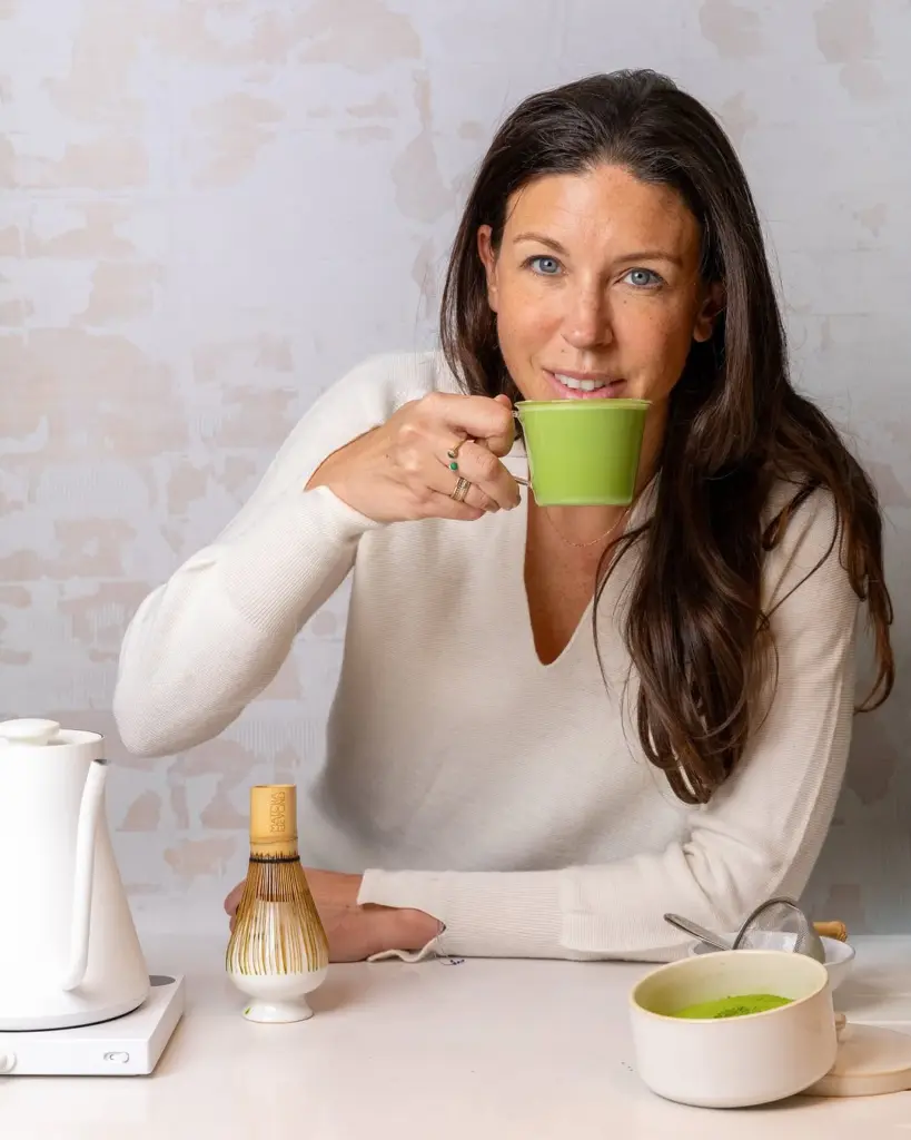 Woman holding a green cup beside matcha preparation tools