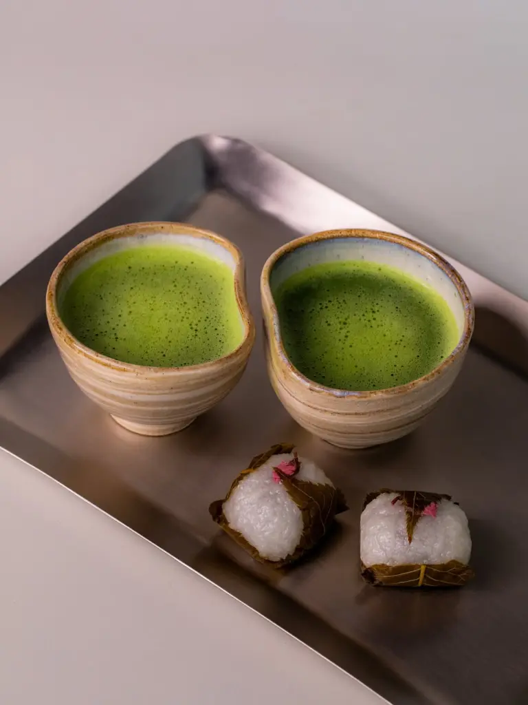 Two ceramic bowls of frothy matcha with leaf-wrapped sweets
