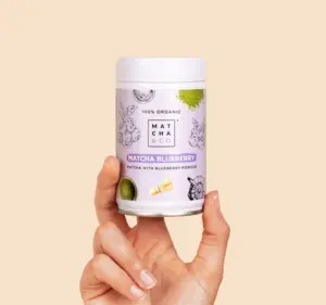 Hand holding a Matcha & CO Matcha Blueberry canister against a pale peach background