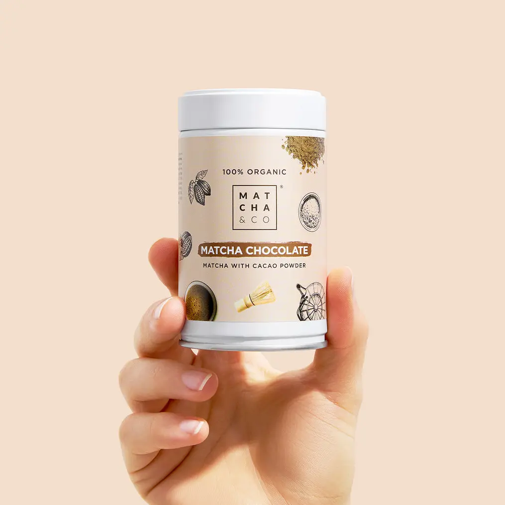 Hand holding a Matcha & CO Matcha Chocolate canister against a pale peach background