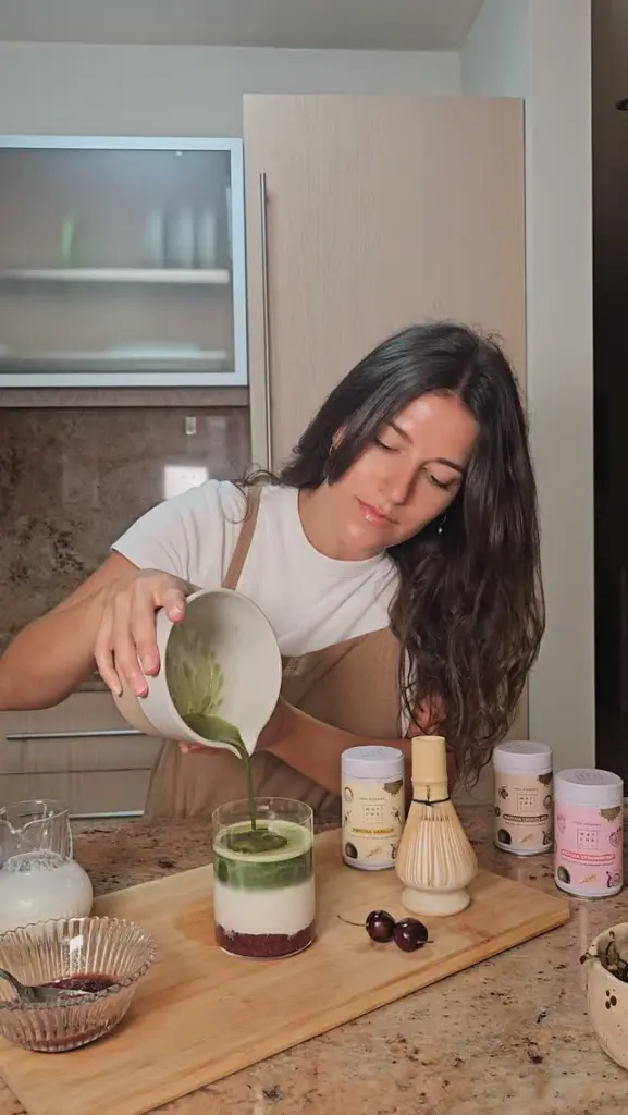 Person pouring a green mixture into a layered drink on a kitchen counter