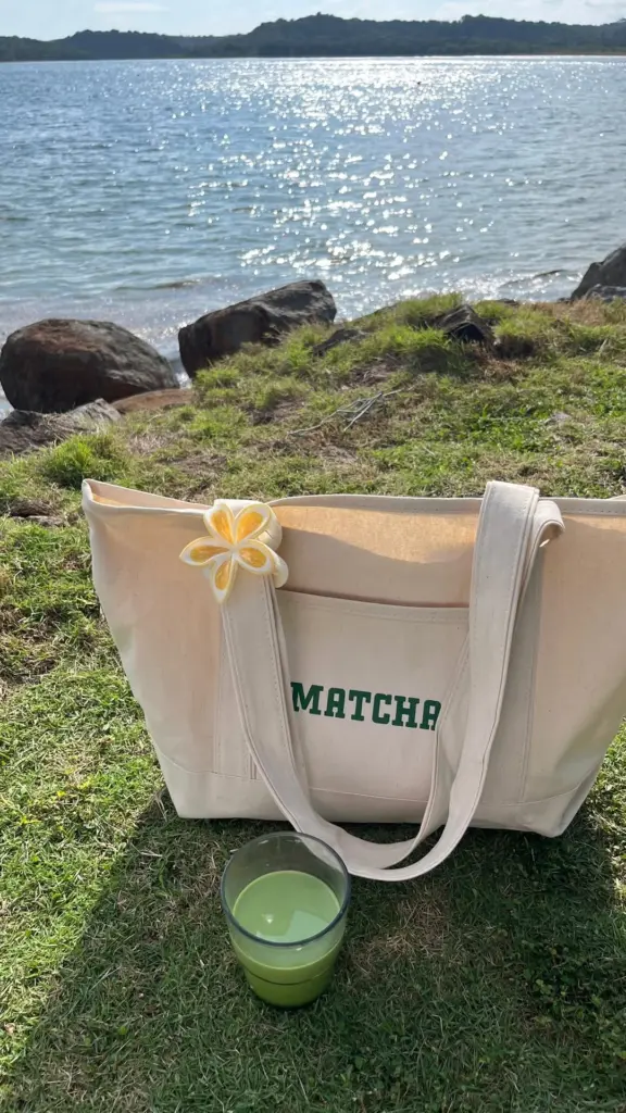 Green drink beside a cream Matcha tote on grass by sunlit water
