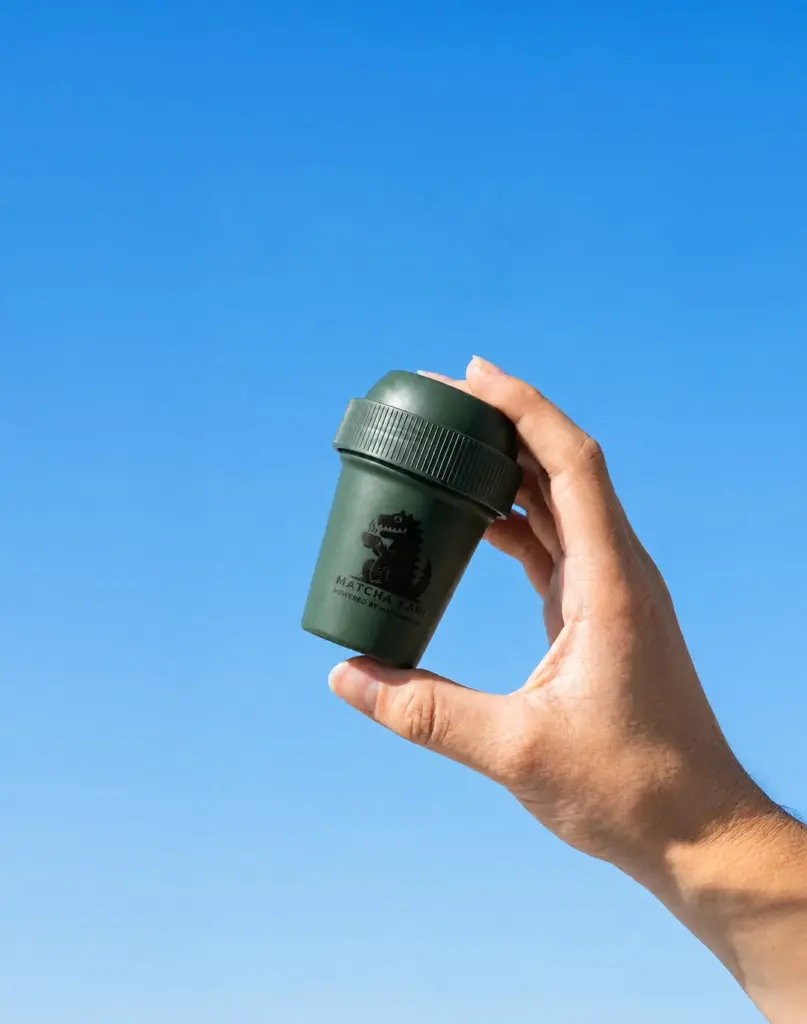 Hand holding a small green matcha shaker against a clear blue sky