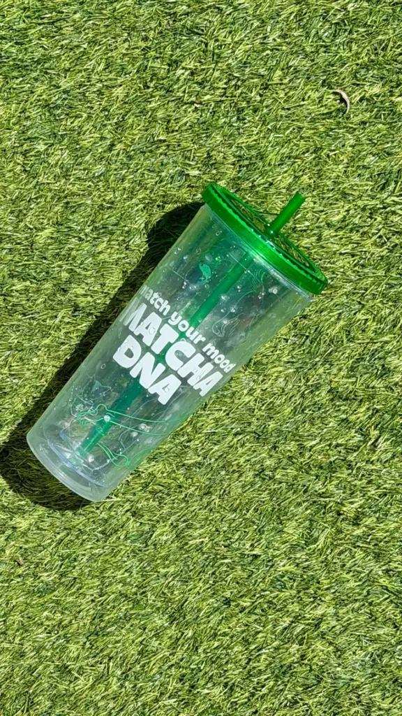 Clear Matcha DNA tumbler with a green lid and straw lying on grass