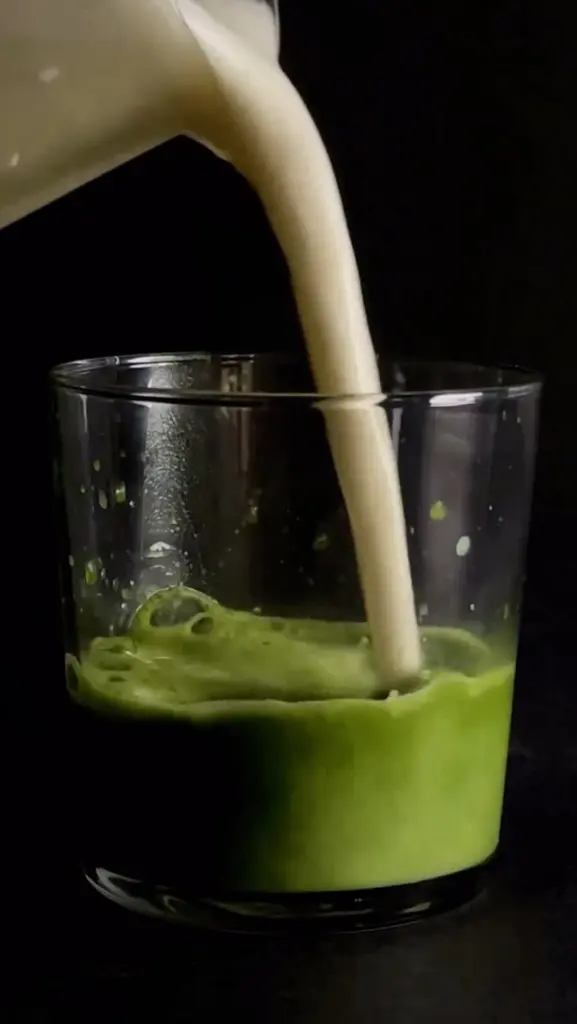 Pale milk pouring into bright green matcha in a clear glass