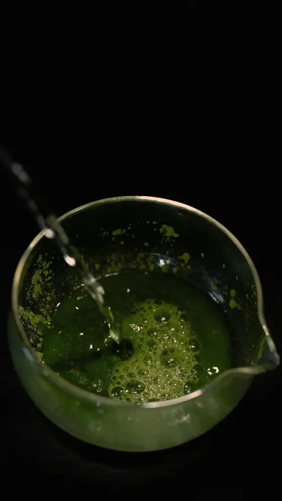 Water pouring into foamy green matcha in a glass pitcher