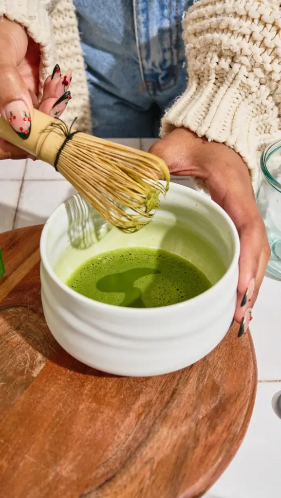 Hands whisking bright green matcha in a white ceramic bowl