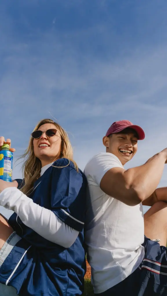 Two smiling friends seated back-to-back under a blue sky, one holding bottled tea
