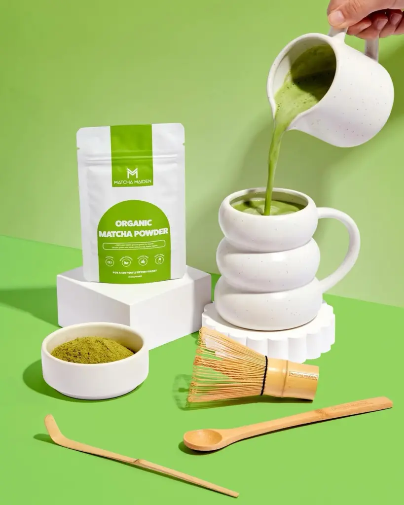 Matcha Maiden powder pouch beside poured matcha, a whisk, spoons, and a bowl of green powder