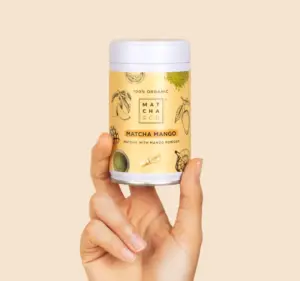 Hand holding a Matcha & CO Matcha Mango canister against a pale beige background