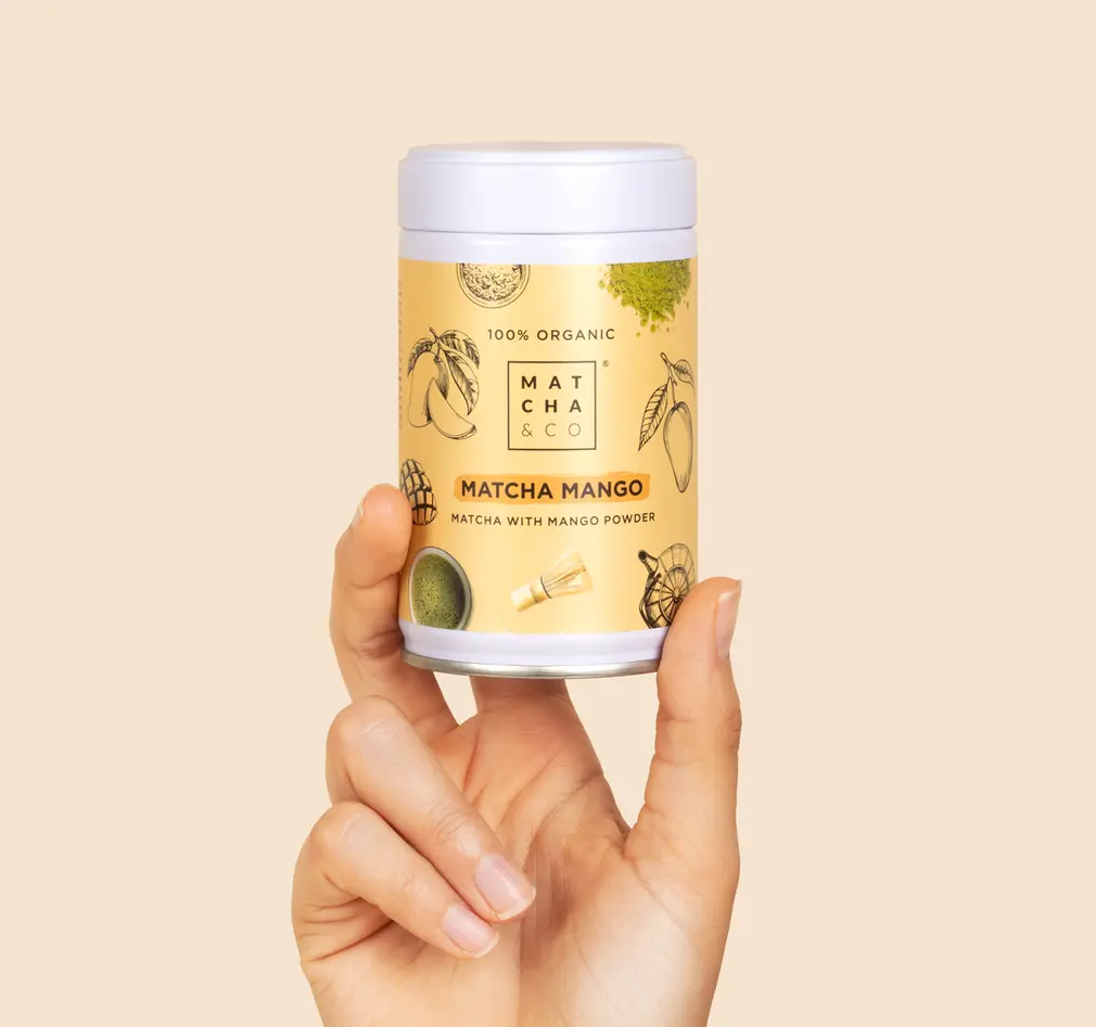 Hand holding a Matcha & CO Matcha Mango canister against a pale beige background