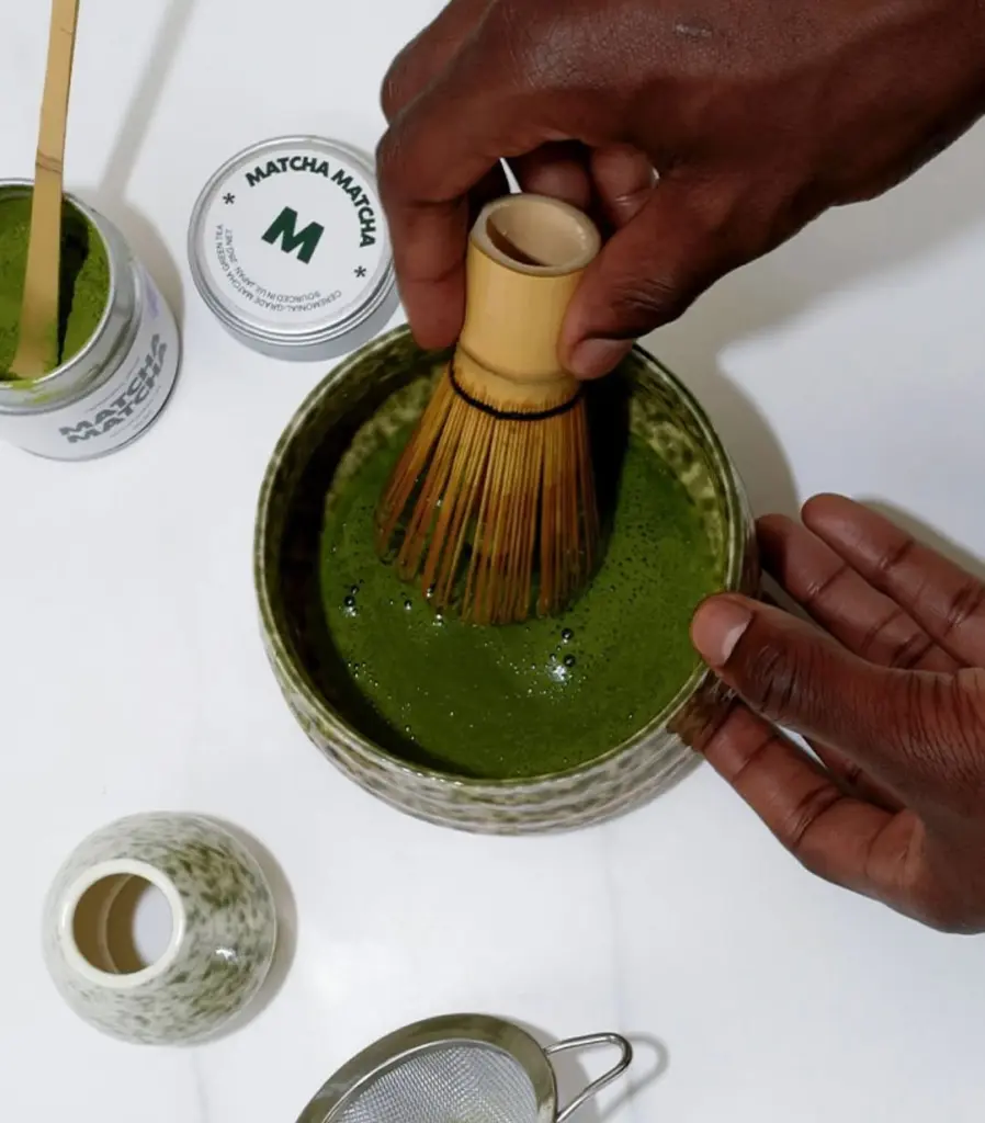 Hands whisking green matcha in a ceramic bowl with a bamboo whisk